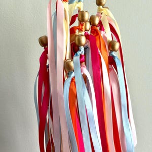 May include: A collection of colorful ribbon wands. Each wand features a gold-painted wooden bead at the top, with long, flowing ribbons in shades of pink, blue, orange, and yellow. The ribbons are tied to the wands.