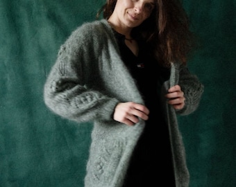 Könnte beinhalten: Eine Frau trägt einen langen, grauen, flauschigen Cardigan mit einem schwarzen Kleid darunter. Der Cardigan hat ein Zopfmuster und ist vorne offen. Die Frau steht vor einem grünen Hintergrund.