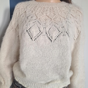 May include: A white knitted sweater with a delicate lace pattern around the neckline. The sweater is made of a soft, fluffy yarn.