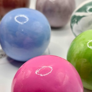 May include: A collection of colorful, glossy spheres in shades of pink, blue, green, purple, and brown. The spheres have a smooth, reflective surface, with a bright highlight on each. The background is a plain white surface.