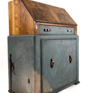 Pode incluir: Uma secret&aacute;ria vintage de madeira com uma frente dobr&aacute;vel e uma &uacute;nica gaveta. A secret&aacute;ria &eacute; pintada em uma cor azul-verde desgastada com uma silhueta de uma pessoa pescando no lado. A secret&aacute;ria tem duas al&ccedil;as de madeira marrom na porta do arm&aacute;rio inferior. O texto "Rugged Refinery Sustainable Home Furniture" &eacute; vis&iacute;vel no canto superior direito da imagem.