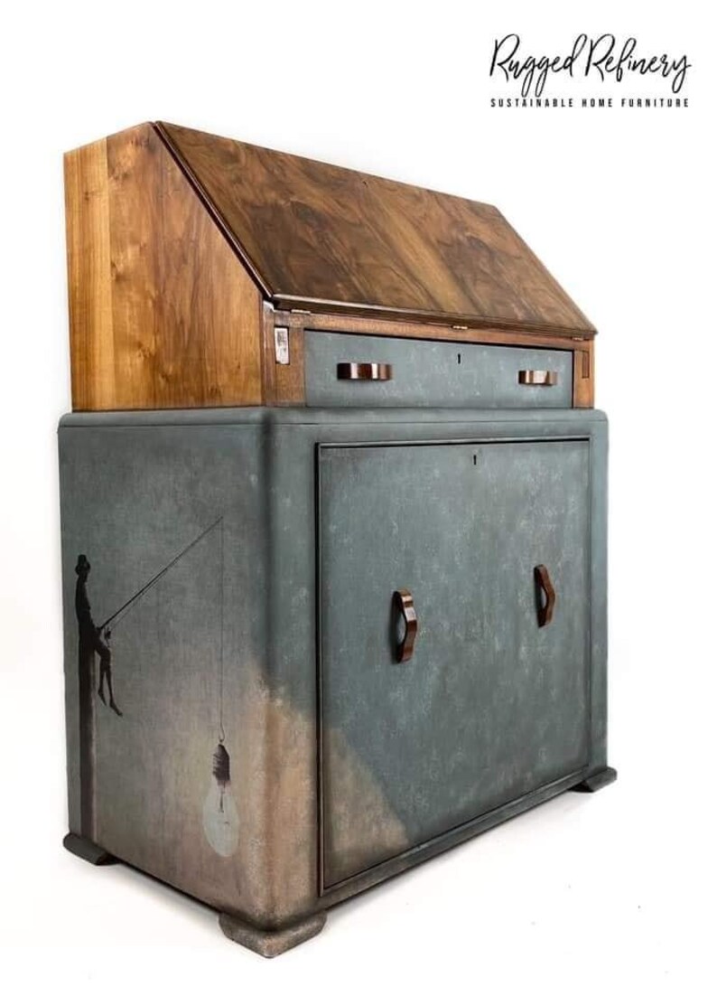 Pode incluir: Uma secret&aacute;ria vintage de madeira com uma frente dobr&aacute;vel e uma &uacute;nica gaveta. A secret&aacute;ria &eacute; pintada em uma cor azul-verde desgastada com uma silhueta de uma pessoa pescando no lado. A secret&aacute;ria tem duas al&ccedil;as de madeira marrom na porta do arm&aacute;rio inferior. O texto "Rugged Refinery Sustainable Home Furniture" &eacute; vis&iacute;vel no canto superior direito da imagem.