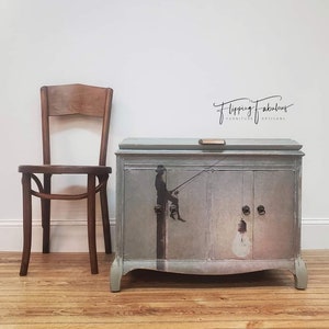 Pode incluir: Um arm&aacute;rio pintado de azul claro com duas portas e uma gaveta. O arm&aacute;rio tem um design caprichoso com uma silhueta de uma pessoa pendurada em uma l&acirc;mpada. O arm&aacute;rio est&aacute; etiquetado como "Flipping Fabulous Furniture Artisans".