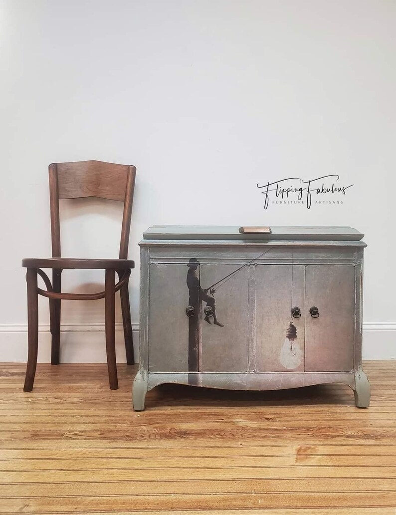 Pode incluir: Um arm&aacute;rio pintado de azul claro com duas portas e uma gaveta. O arm&aacute;rio tem um design caprichoso com uma silhueta de uma pessoa pendurada em uma l&acirc;mpada. O arm&aacute;rio est&aacute; etiquetado como "Flipping Fabulous Furniture Artisans".