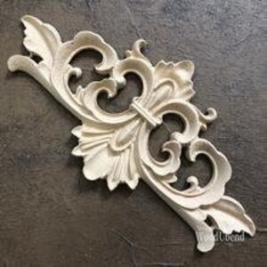 Pode incluir: Uma escultura decorativa em madeira creme, ornamentada com um design floral e de rolagem simétrico. A escultura apresenta folhas detalhadas e padrões em espiral, adequada para decoração de móveis ou paredes.