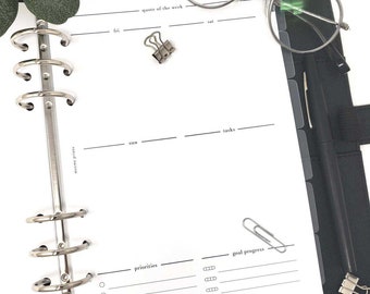 May include: A white weekly planner with a black binder, a silver paper clip, and a black pen. The planner has sections for "quote of the week", "fri", "sat", "sun", "tasks", "priorities", and "goal progress".