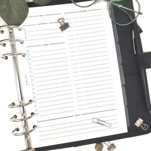 May include: A white planner insert with sections for date, schedule, priorities, tasks, and notes. The planner is held in a black binder with silver rings. A silver binder clip and paper clip are attached to the page.