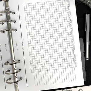 May include: A black binder with a white page open to a habit tracker. The tracker has a grid with numbered columns for each day of the month. The page also has a section for notes.