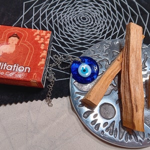 May include: A silver tray containing two brown sticks of Palo Santo wood, a blue glass evil eye necklace, and a small brown crystal. The tray is on a black fabric background with a white geometric pattern.