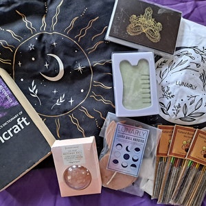 May include: A collection of witchy items including a book titled "Cassell's Dictionary of Witchcraft", a black tapestry with a gold sun and moon design, a white tote bag with a black floral design and the word "LUNARLY", a green gua sha tool, a wooden moon phase wall hanging, and a pack of incense sticks.