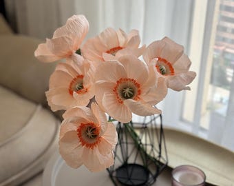 Könnte beinhalten: Ein Strauß pfirsichfarbener Papier-Mohnblumen mit orangefarbenen und grünen Zentren in einer schwarzen geometrischen Vase. Die Blumen stehen auf einem weißen Tisch neben einer brennenden Kerze und einem goldenen Tablett. Der Hintergrund umfasst ein Fenster und ein beiges Sofa.