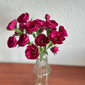 May include: A bouquet of deep magenta paper flowers in a clear glass bottle vase. The flowers have multiple layers of petals and green stems. The vase sits on a brown wooden surface against a white wall.