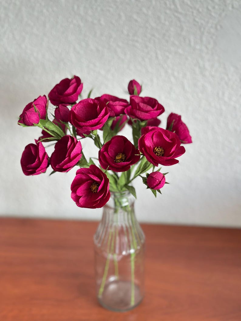 May include: A bouquet of deep magenta paper flowers in a clear glass bottle vase. The flowers have multiple layers of petals and green stems. The vase sits on a brown wooden surface against a white wall.
