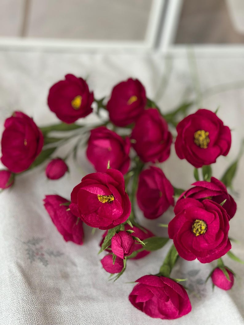 May include: A collection of deep magenta paper flowers with yellow centers and green stems. The flowers are in various stages of bloom, from tightly closed buds to fully open blossoms. The arrangement is set against a white, patterned background.