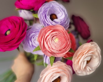 May include: A bouquet of paper flowers in shades of pink, purple, and peach. The flowers have detailed, layered petals and green paper leaves. The bouquet is held against a blurred background.