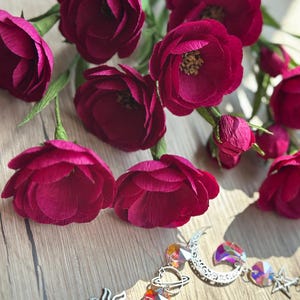 May include: Burgundy paper flowers with green stems are arranged on a wooden surface. A decorative sun, moon, and star crystal prism hangs below the flowers. The prism features iridescent crystals and silver-toned metal charms.