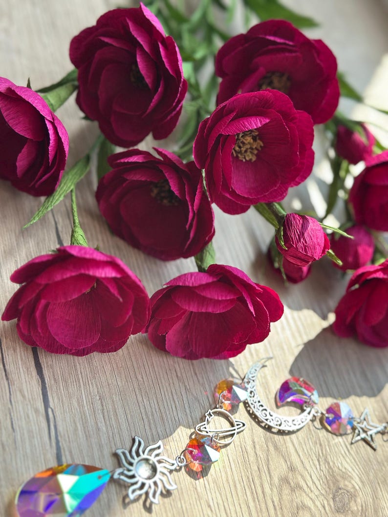 May include: Burgundy paper flowers with green stems are arranged on a wooden surface. A decorative sun, moon, and star crystal prism hangs below the flowers. The prism features iridescent crystals and silver-toned metal charms.