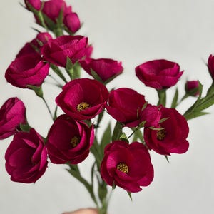 May include: A bouquet of deep magenta paper flowers with yellow centers and green stems and leaves. The flowers are in full bloom, with multiple petals visible. The background is a plain white.