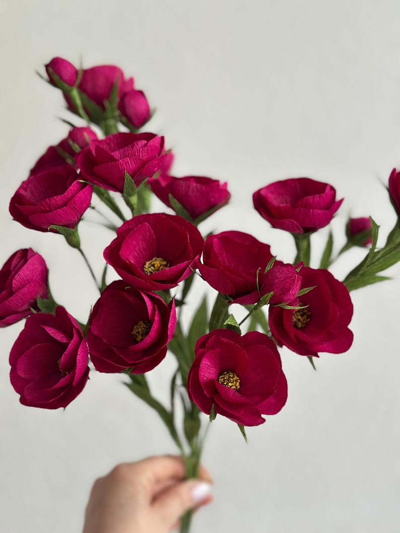 May include: A bouquet of deep magenta paper flowers with yellow centers and green stems and leaves. The flowers are in full bloom, with multiple petals visible. The background is a plain white.