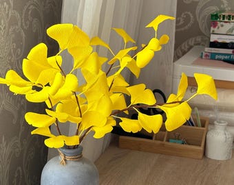 May include: A gray ceramic vase with a rounded base and a narrow neck, tied with a piece of twine. Inside the vase are several branches featuring bright yellow, fan-shaped ginkgo leaves made of paper, creating a vibrant and textured display.