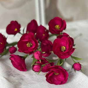 May include: A cluster of deep magenta paper flowers with yellow centers and green stems. The flowers are in various stages of bloom, with some tightly closed buds. The arrangement is set against a soft, white background.