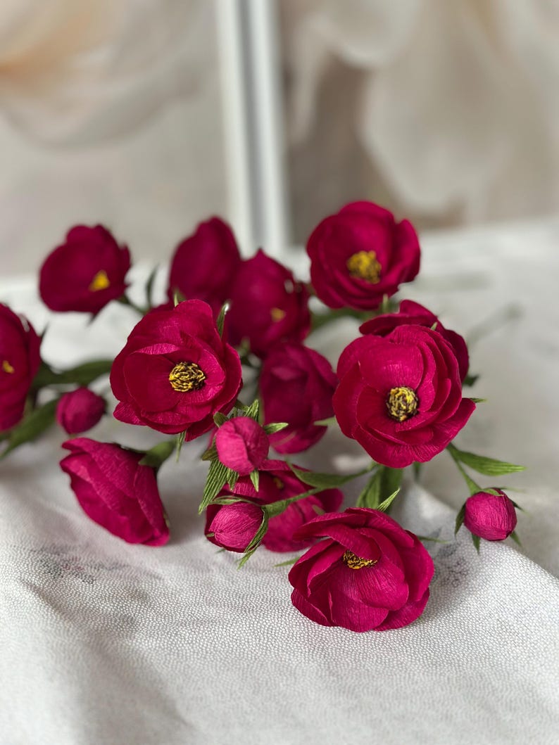 May include: A cluster of deep magenta paper flowers with yellow centers and green stems. The flowers are in various stages of bloom, with some tightly closed buds. The arrangement is set against a soft, white background.
