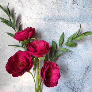 May include: A bouquet of handmade paper flowers with deep magenta petals and green leaves. The flowers have a textured appearance and are arranged against a light blue and gray background.
