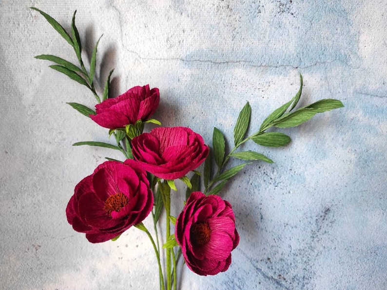May include: A bouquet of handmade paper flowers with deep magenta petals and green leaves. The flowers have a textured appearance and are arranged against a light blue and gray background.