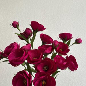 May include: A bouquet of deep magenta paper flowers with green stems and leaves. The flowers are in various stages of bloom, with some tightly closed buds and others fully open, revealing yellow centers. The arrangement is held against a white background.