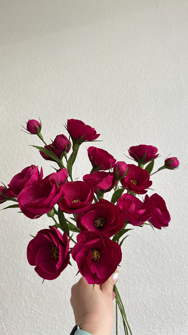 May include: A bouquet of deep magenta paper flowers with green stems and leaves. The flowers are in various stages of bloom, with some tightly closed buds and others fully open, revealing yellow centers. The arrangement is held against a white background.