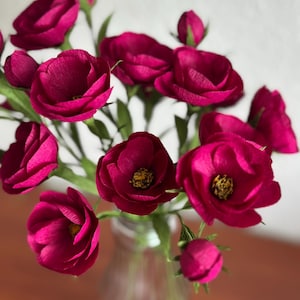 May include: A bouquet of deep magenta paper flowers in a clear glass vase. The flowers have multiple layers of petals and yellow centers. Green stems and leaves are visible. The vase sits on a brown surface.