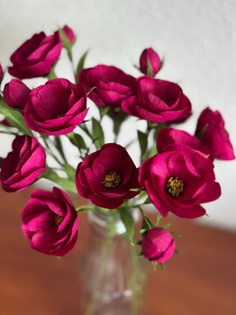 May include: A bouquet of deep magenta paper flowers in a clear glass vase. The flowers have multiple layers of petals and yellow centers. Green stems and leaves are visible. The vase sits on a brown surface.