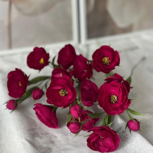 May include: A collection of deep magenta paper flowers with yellow centers and green stems. The flowers are in various stages of bloom, with some tightly closed buds. The arrangement is set against a soft, textured background.