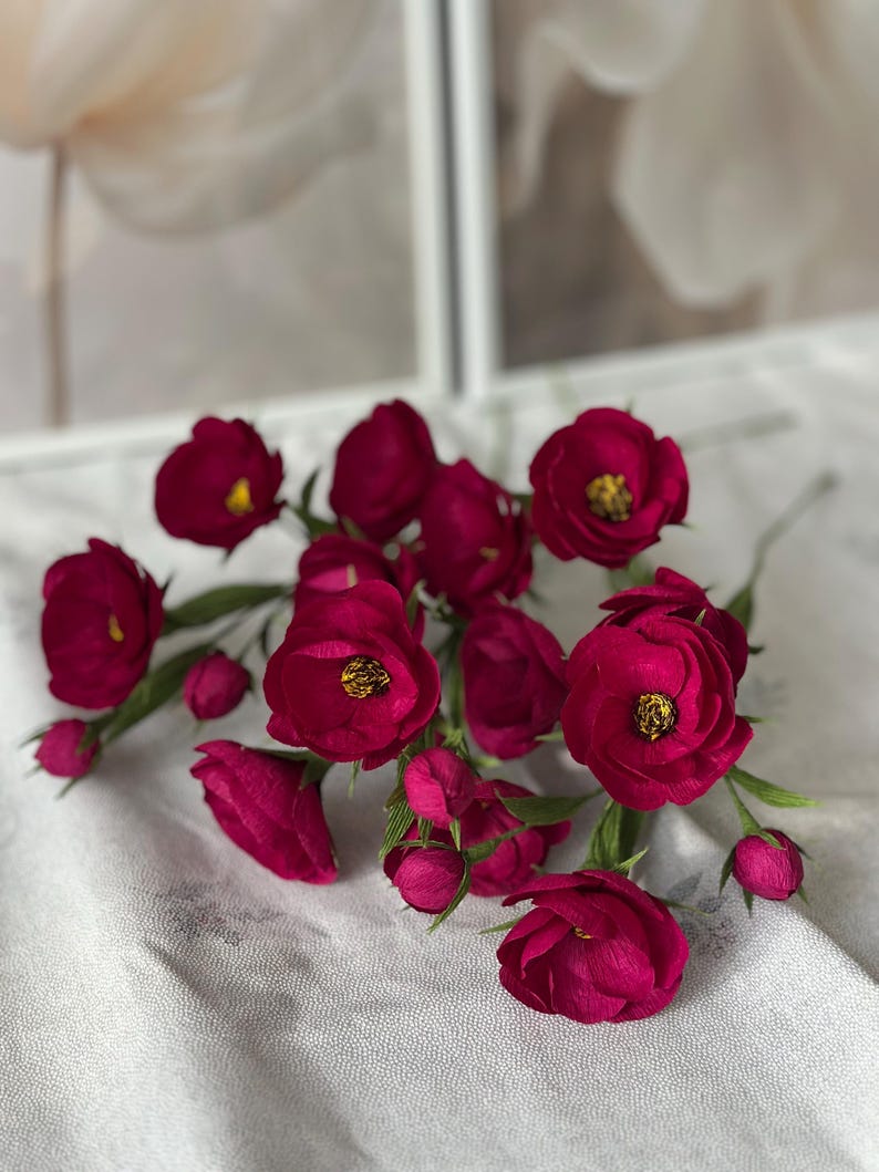 May include: A collection of deep magenta paper flowers with yellow centers and green stems. The flowers are in various stages of bloom, with some tightly closed buds. The arrangement is set against a soft, textured background.