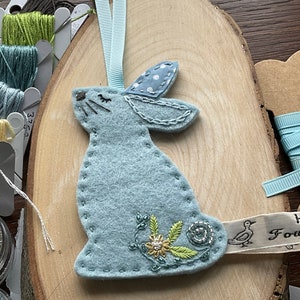 May include: A pale blue felt bunny decoration with embroidered details and a light blue ribbon. The bunny features a floral design and is stitched with white thread. It rests on a wooden slice, with spools of thread and beads visible in the background.