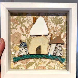 May include: Framed artwork depicting a village scene made from ceramic pieces. The piece includes a white triangular roof and houses with tan and brown roofs, set against a floral patterned background. A green rope-like element forms the base of the artwork.