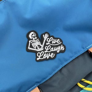 Puede incluir: Una mochila azul con un parche bordado blanco que dice "Live Laugh Love" y un gráfico de esqueleto.