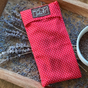 May include: A red, polka-dotted pouch with a label that reads "Made by Julie" rests on a wooden tray with dried lavender and a bowl of seeds. The pouch is rectangular and folded over.