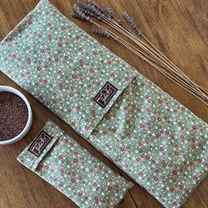 May include: A green floral-patterned heat pack and eye pillow set with a small bowl of seeds and dried lavender sprigs. The heat pack and eye pillow have a green background with small red and white flowers. The set has a brown label that says "Julie".