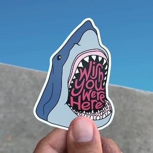 May include: A blue and white shark sticker with an open mouth. The text inside the mouth reads "Wish you were here".