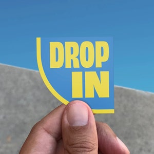 Puede incluir: Una pegatina azul y amarilla con el texto "DROP IN" en ella.