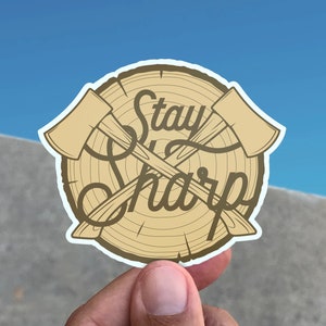 Op de afbeelding: Een ronde sticker met een houtnerfdesign en twee gekruiste bijlen. De tekst "Stay Sharp" is geschreven in een gestileerd lettertype.