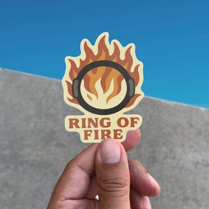Könnte beinhalten: Ein Aufkleber mit einem schwarzen Ring mit Kopfhörern, umgeben von Flammen. Der Text "RING OF FIRE" ist in einem braunen Banner darunter zu sehen. Der Aufkleber wird vor einem blauen Himmel und einem grauen Betonhintergrund gehalten.
