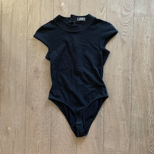 May include: Black bodysuit with short sleeves and a mock neck. The bodysuit has a snap closure at the bottom and the label "Gianni Versace" is visible on the back of the neck.