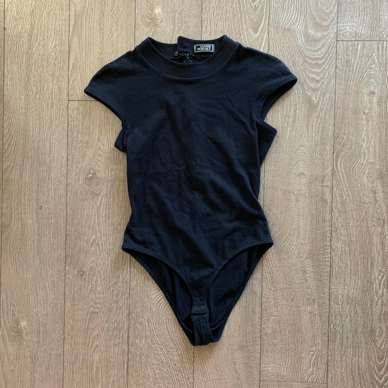 May include: Black bodysuit with short sleeves and a mock neck. The bodysuit has a snap closure at the bottom and the label "Gianni Versace" is visible on the back of the neck.