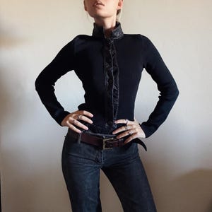 May include: Black long-sleeved top with a high collar and leather trim. The top is buttoned down the front and is paired with dark denim jeans and a brown leather belt with a gold buckle. The outfit is a classic, stylish look.
