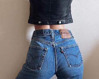 May include: A black leather crop top paired with blue denim jeans. The jeans have a classic five-pocket design with a leather patch and red tab detail. The jeans are a medium wash.