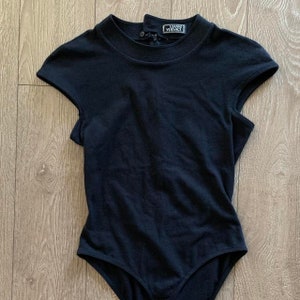 May include: Black bodysuit with short sleeves and a mock neck. The bodysuit has a snap closure at the back and the label "GIANNI VERSACE" is printed on the back of the neck.