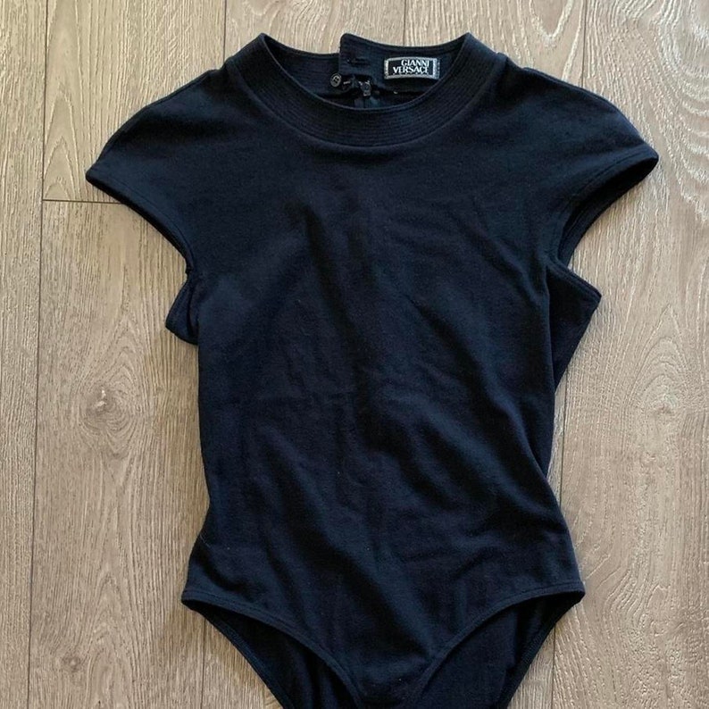 May include: Black bodysuit with short sleeves and a mock neck. The bodysuit has a snap closure at the back and the label "GIANNI VERSACE" is printed on the back of the neck.