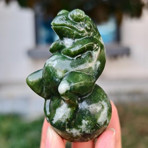 Op de afbeelding: Een groene jadesculptuur van een kikker met witte marmering. De kikker zit op een ronde basis en heeft zijn poten gekruist.